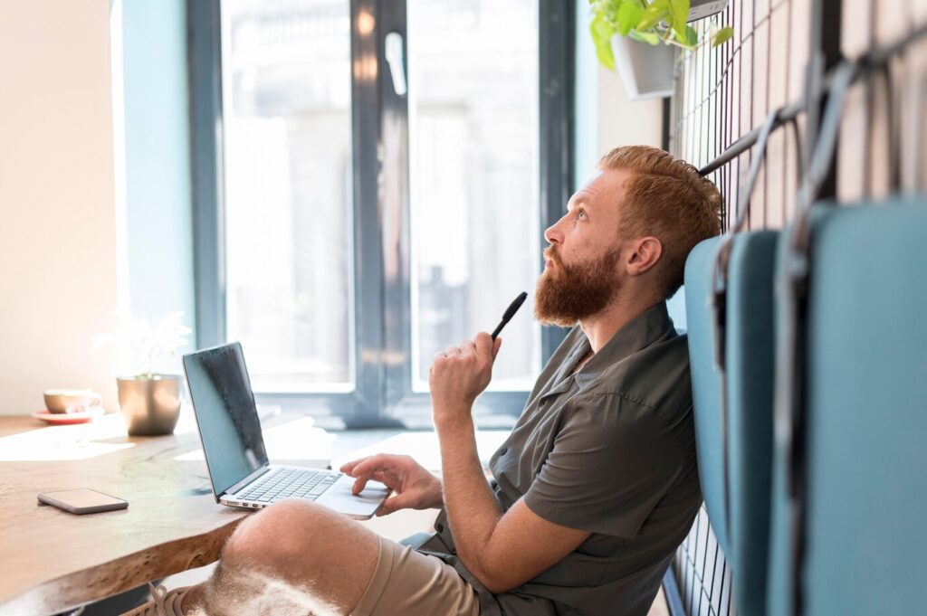 person sitting at a table with a laptop, thoughtfully holding a pen