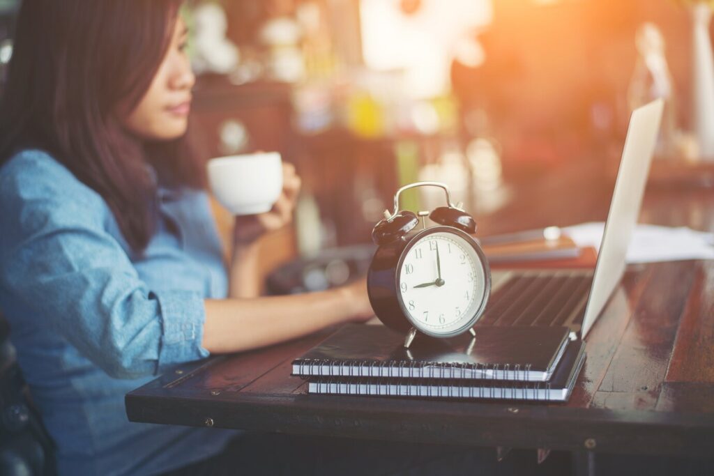 person sits at a wooden table with a laptop, an alarm clock