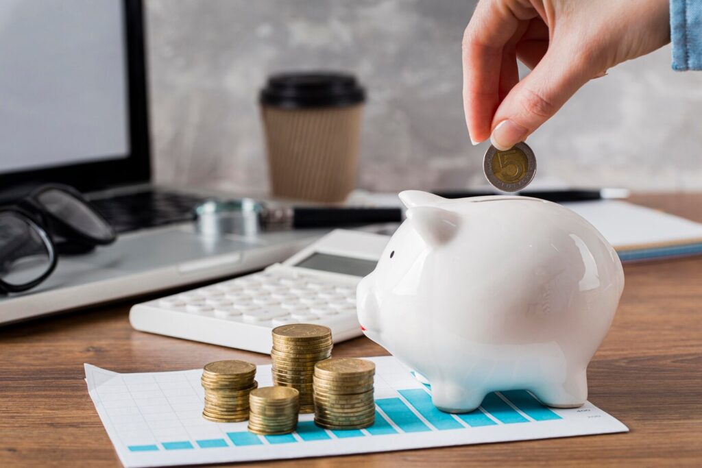 hand drops a coin into a white piggy bank on a desk with coins