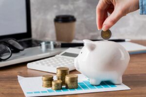 hand drops a coin into a white piggy bank on a desk with coins