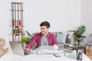 Young man studying with laptop and books at a white desk in a comfortable living room setting.