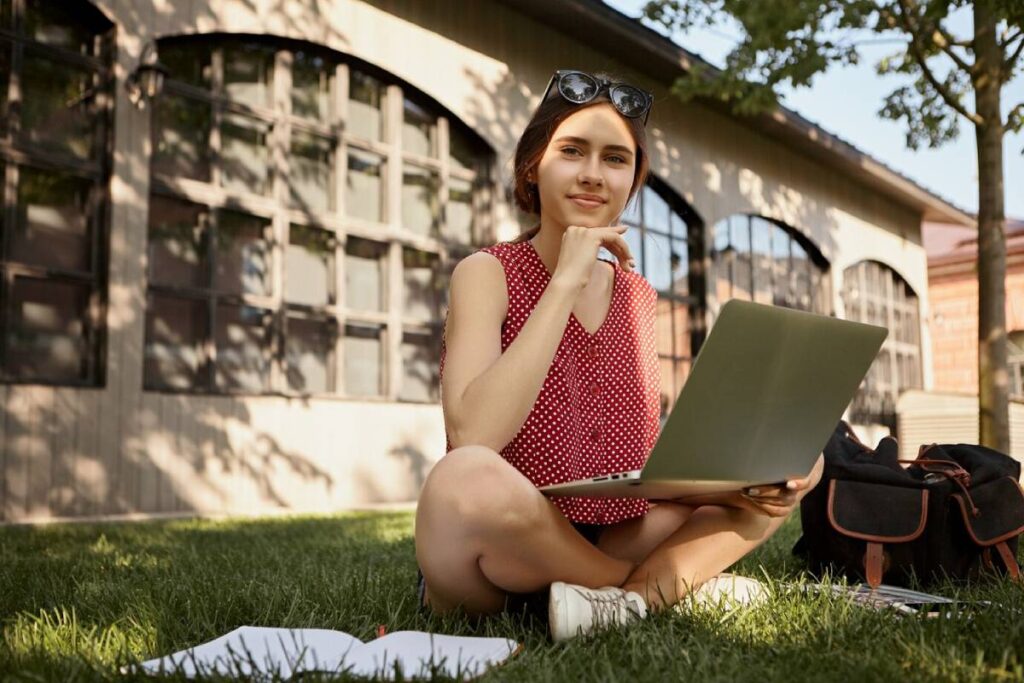 Young woman using a laptop while sitting on grass on a sunny day, with a building in the background.