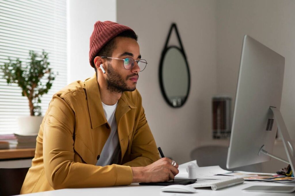 A man in a yellow jacket and red beanie working intently at a computer desk.