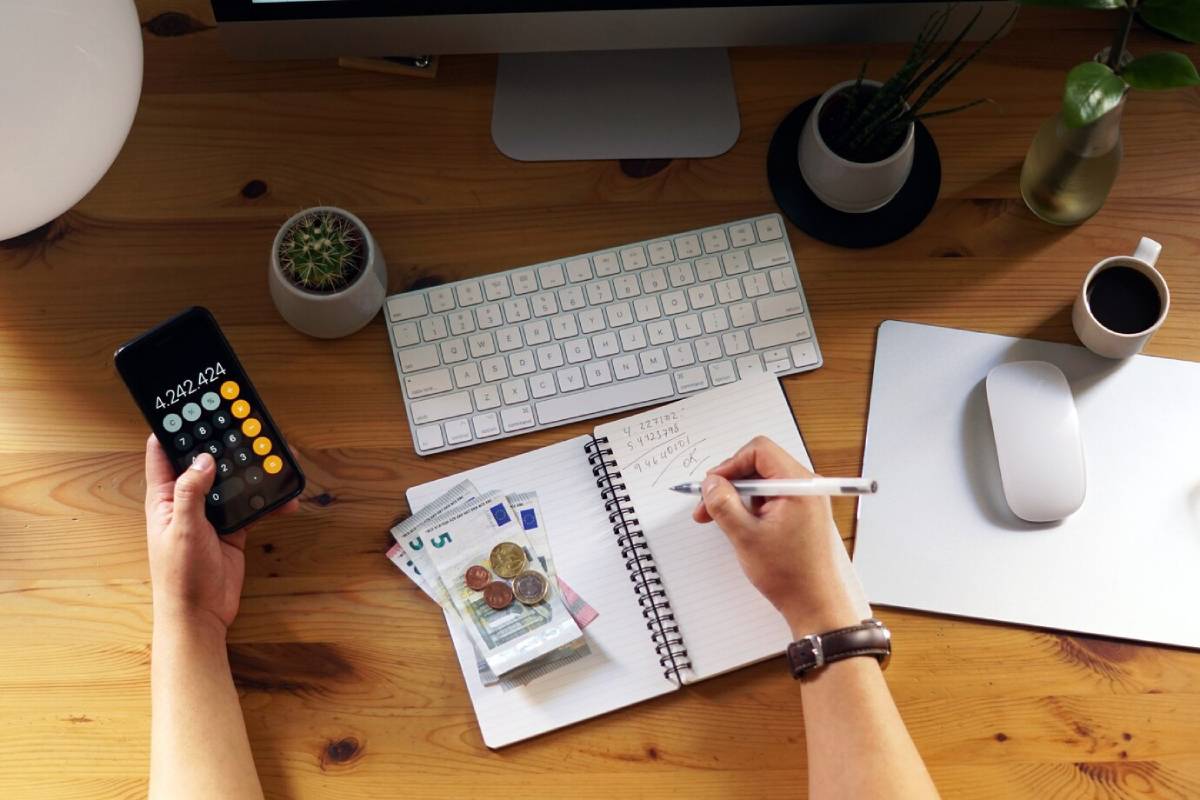 Person calculating finances on a calculator at a wooden desk with a computer, notepad, and money. 