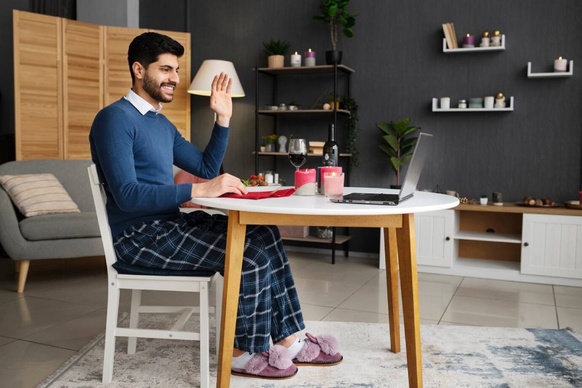 Man in cosy home attire using a laptop and waving, seated at a dining table in a stylish room. 