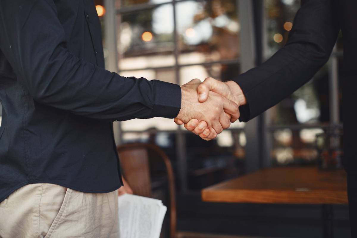 Two individuals shaking hands, one in a black blazer, in front of a café.