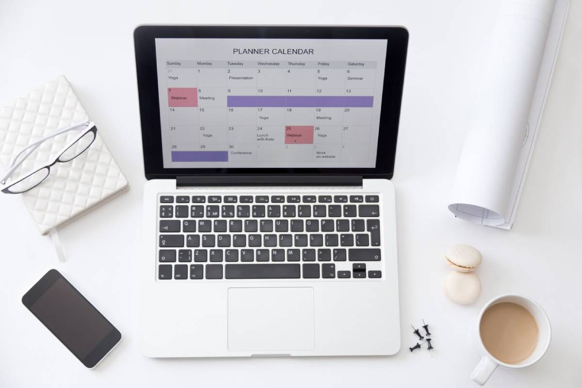 A laptop displaying a calendar, surrounded by a notebook, glasses, smartphone, coffee cup, macarons, and rolled-up papers on a white desk.