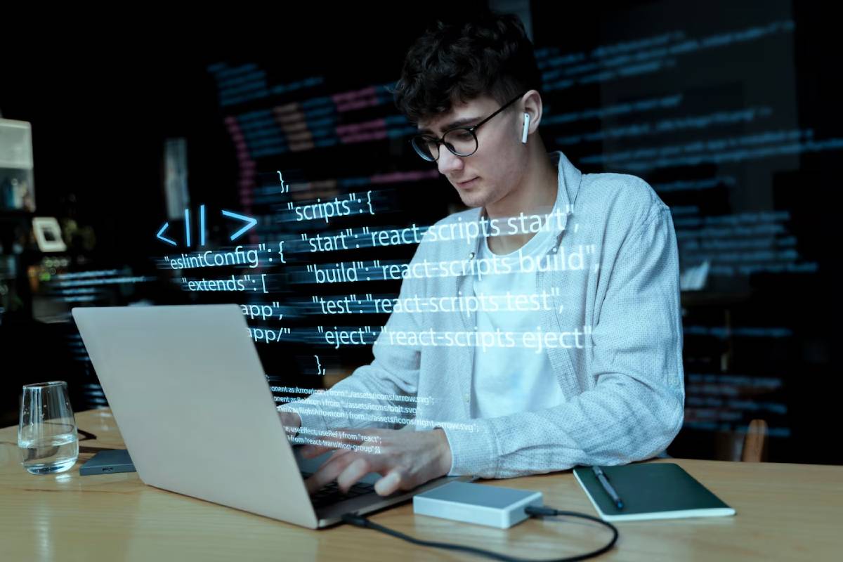 Young man coding on laptop with digital coding graphics overlaid, in a dimly lit office setting.