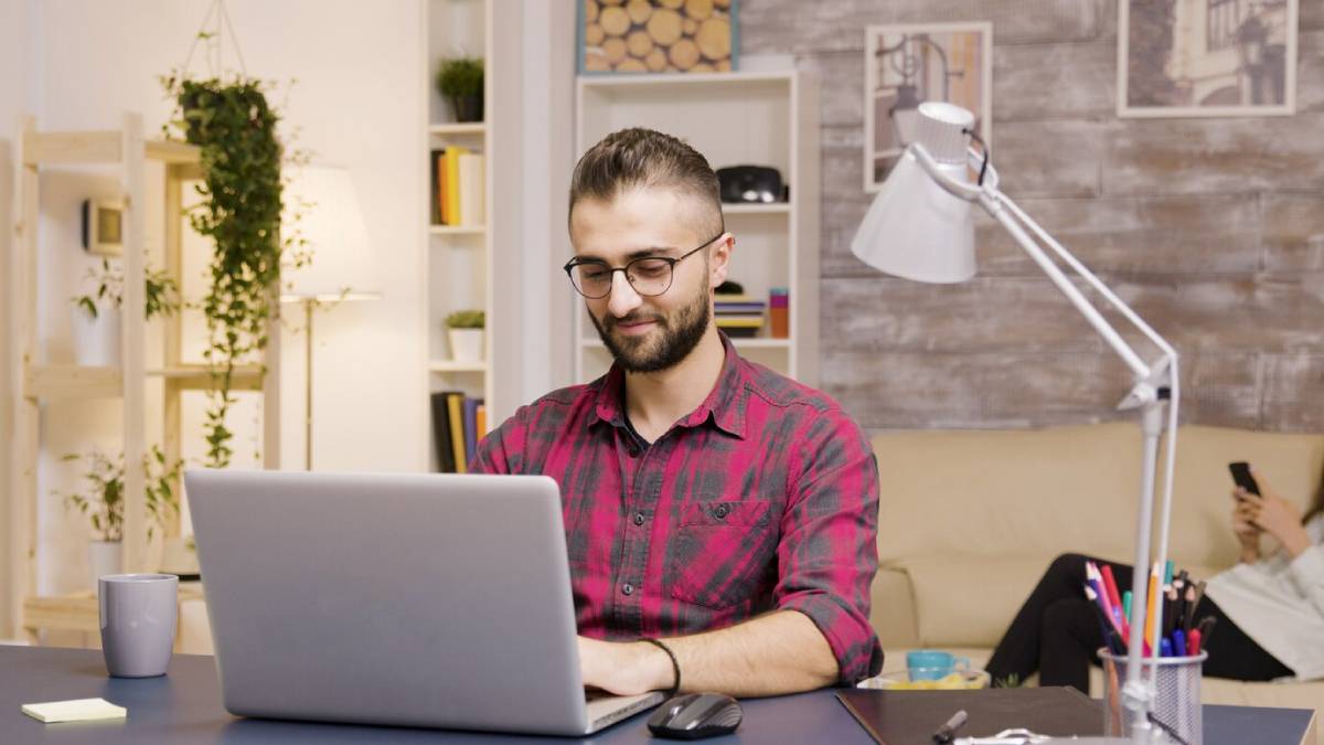  Man with glasses smiling while working on laptop in a well-lit, cozy home office.