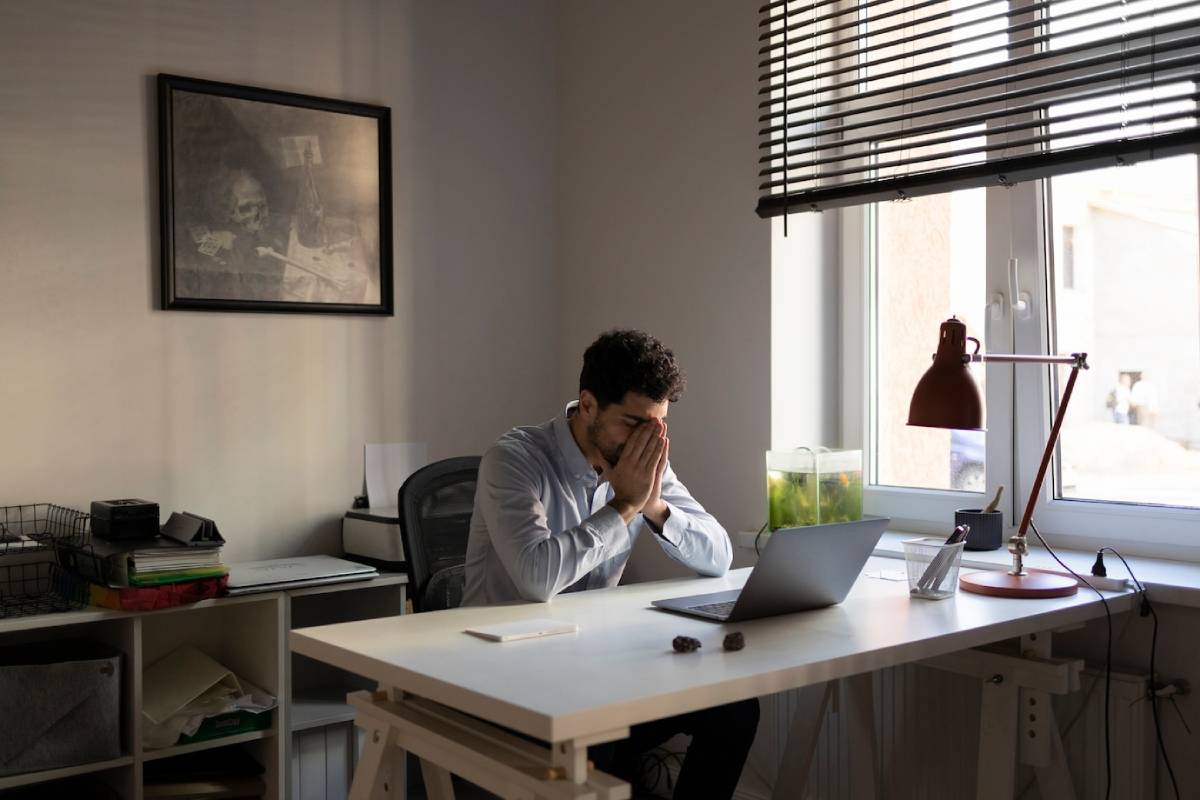 Man sitting at a desk in a sunlit office, looking stressed with hands covering face. 