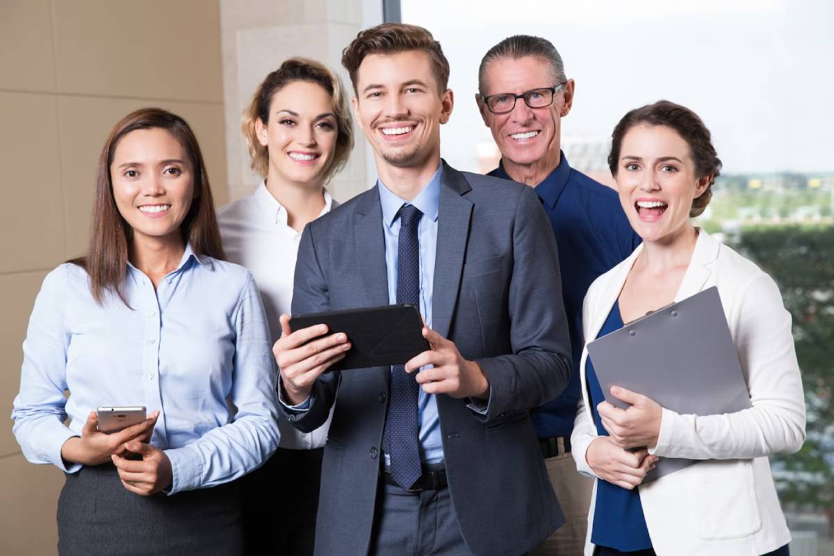 Five office colleagues smile, holding phones and tablets, in a modern business environment. 